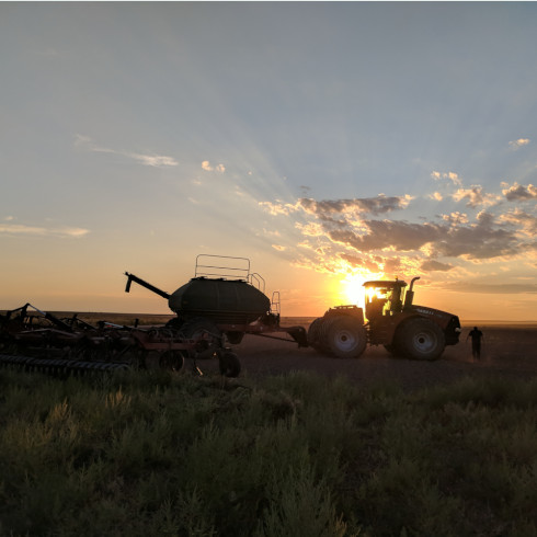 a tractor hooked to planting equipment sits in front of the setting sun as the silohuette of a person walks to the left of it
