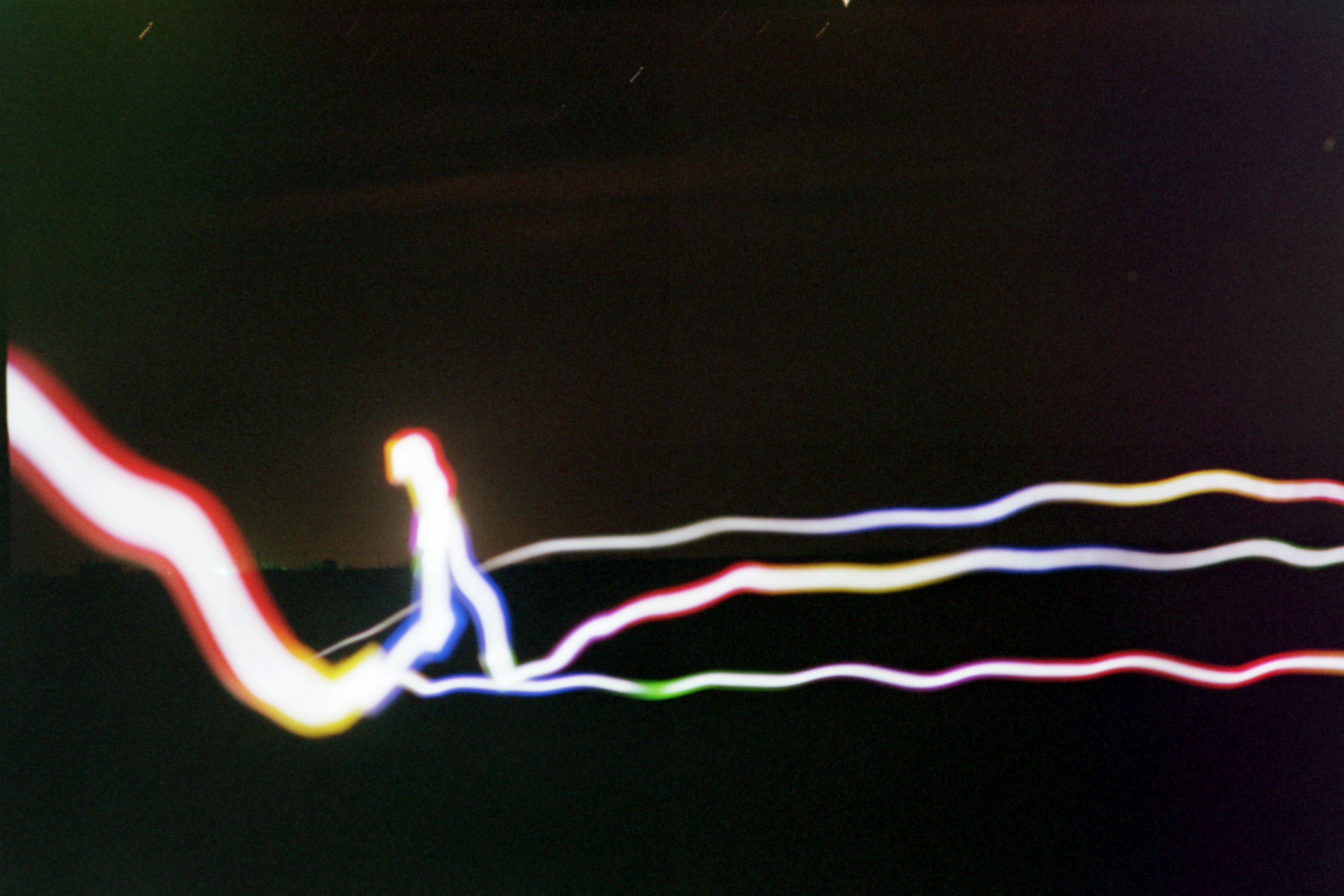 colorful streaks of light go across a mostly black background where just the hint of light over the horizon backlights some trees