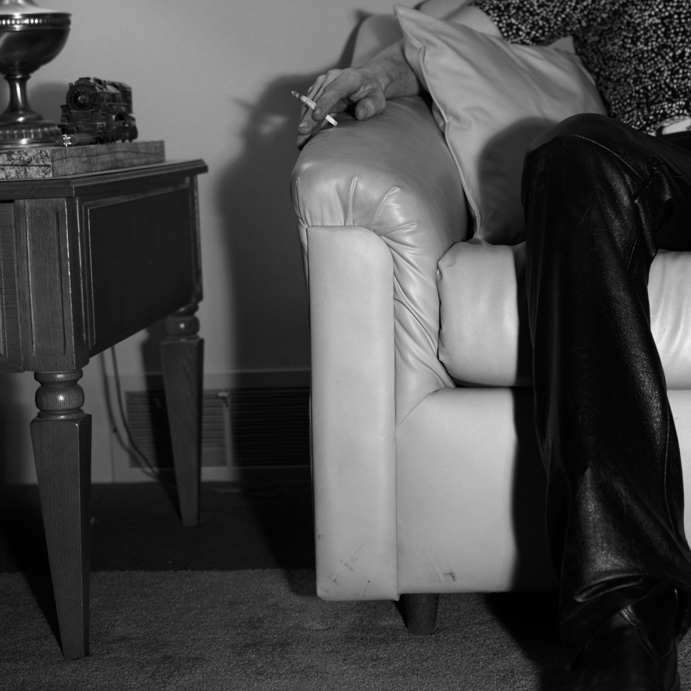 an oddly cropped photo of a person sitting on a couch wearing leather pants and smoking a hand rolled cigarette