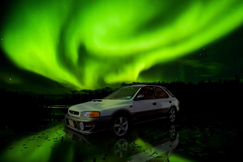 a white subaru impreza hatchback sits in shallow water under a glorious display of aurora borealis, the reflections of which engulf the car